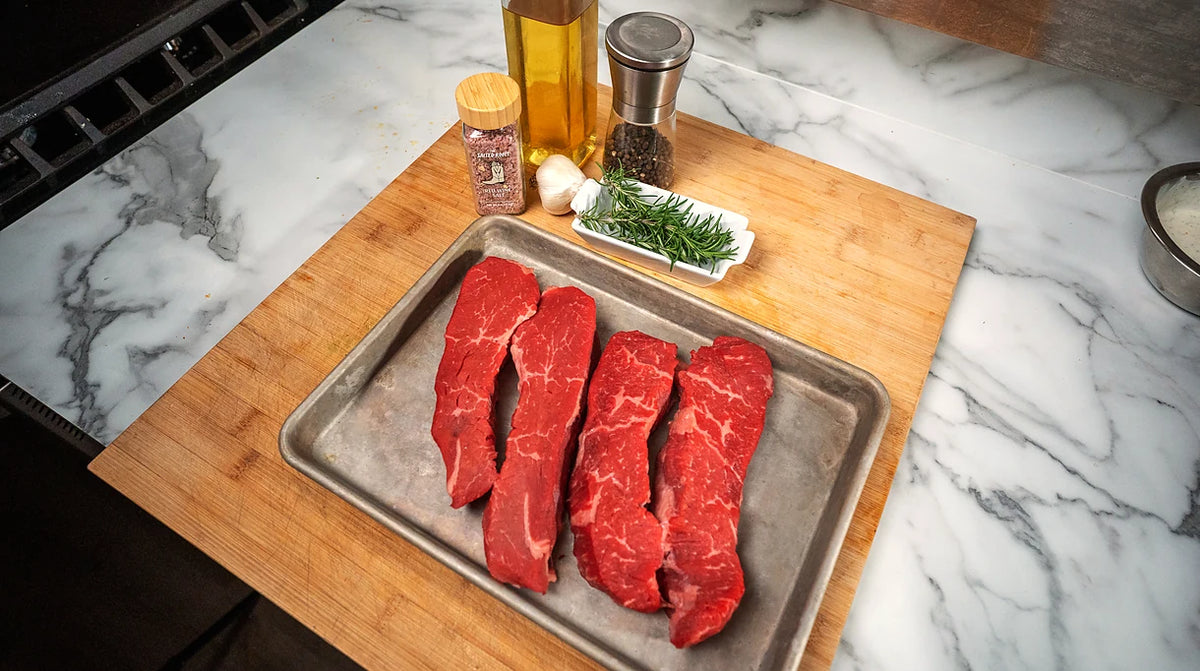 Tri-tip steaks on a sheet pan beside The Salted Roots Red Wine Salt, garlic, rosemary and olive oil