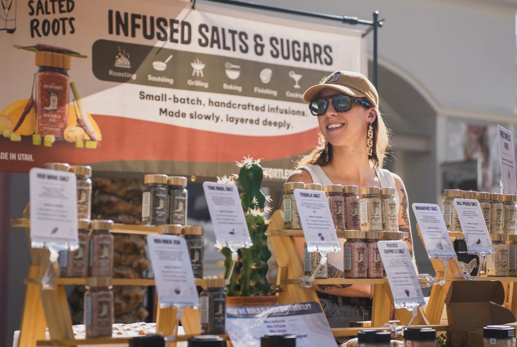 Woman standing behind a display of 'The Salted Roots' infused salts and sugars at an outdoor market.